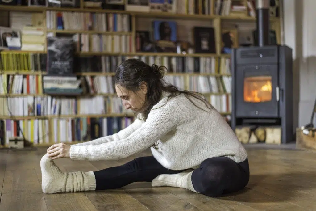 Femme en flexion avant assise, jambes tendues, près d'un poêle à bois en hiver
