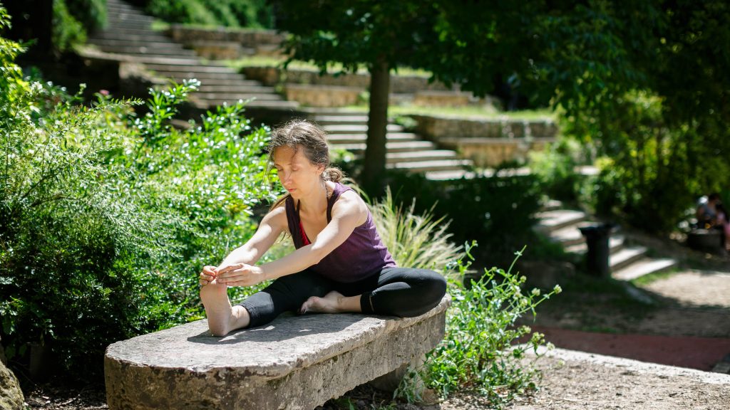 Femme en flexion avant assise sur un banc de pierre, dans un jardin verdoyant